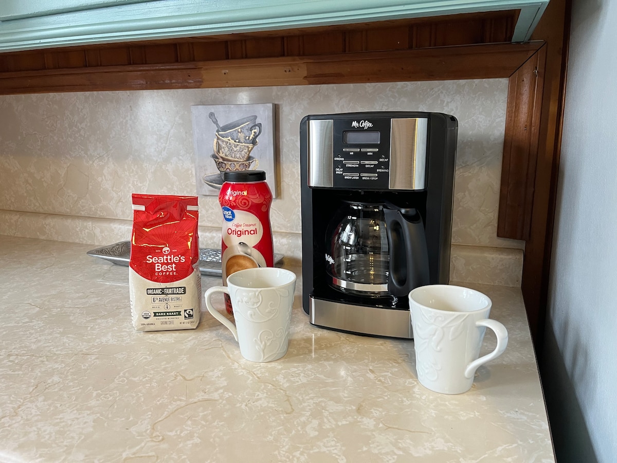 A coffee station is set on a light-colored countertop, featuring a coffee maker, two white mugs, a bag of Seattle's Best coffee, and a container of coffee creamer. A small decorative print is positioned in the background.