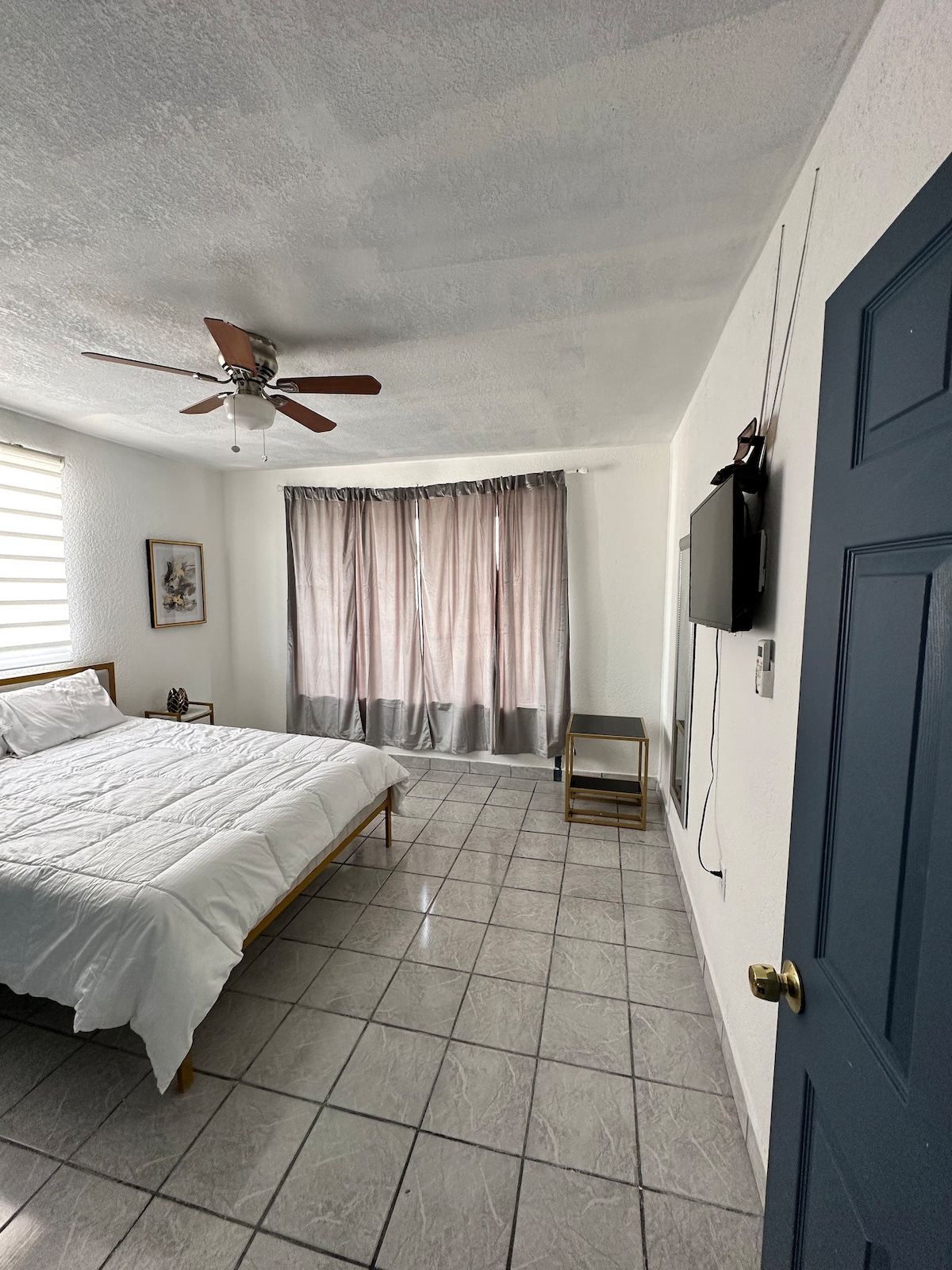 A tidy bedroom features a spacious bed with crisp white linens. A ceiling fan circulates air, while curtains frame the window, allowing soft light to enter. A small nightstand is present beside the bed, and a television is mounted on the wall opposite.