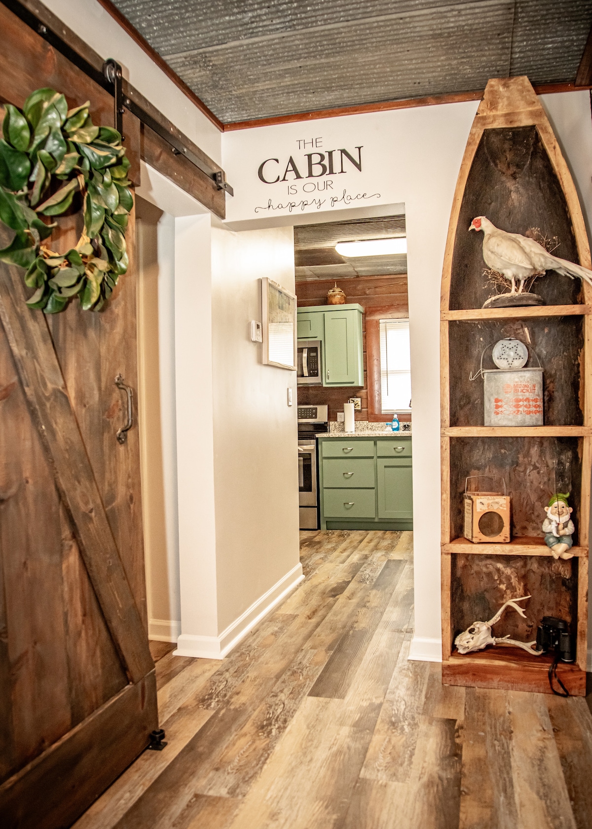 A rustic entryway features a sliding wooden door that opens to a modern kitchen, complemented by green cabinets. Natural wood flooring extends throughout the space. Shelving made from wood displays decorative items and a small bird, enhancing the cabin's charm.