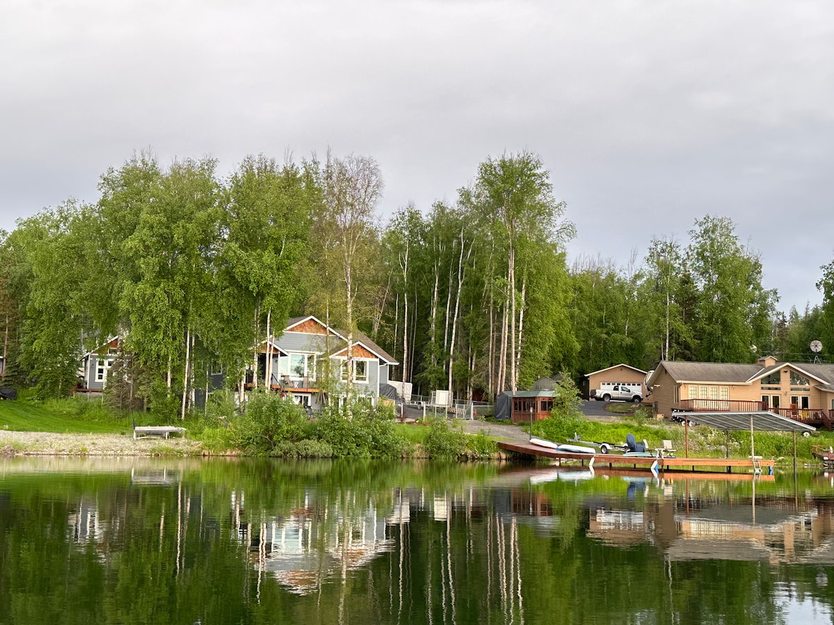 A serene lakeside view captures the guest house and neighboring properties surrounded by lush greenery. The calm water reflects the structures and surrounding trees, enhancing the peaceful atmosphere. A dock with boats is visible, inviting opportunities for fishing and boating.