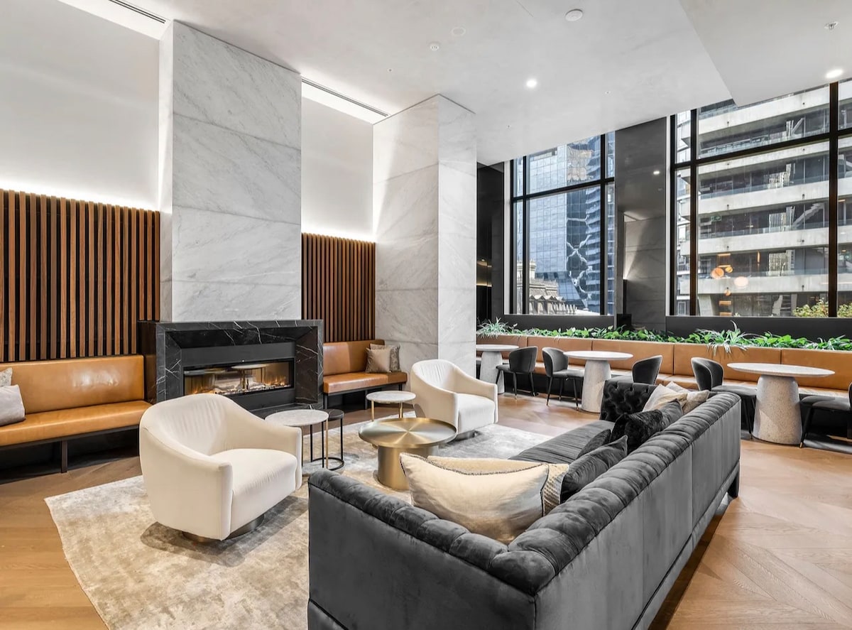 A modern lobby area is presented with large windows allowing natural light to enter. Comfortable seating arrangements include a plush sofa and stylish armchairs. A sleek fireplace is centered against a marble wall, complementing the wooden paneling and greenery that decorates the space.