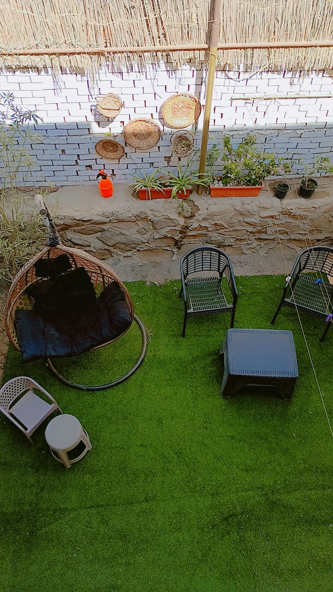 An outdoor seating area is presented on a green lawn, featuring a hanging chair, four black chairs, and a low table. Decorative baskets are mounted on the white brick wall, while potted plants add a touch of greenery to the space. A tranquil atmosphere is suggested.