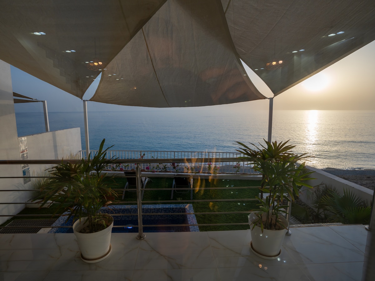 A view from a covered terrace features potted plants and a sparkling pool overlooking the sea. The horizon is framed by the setting sun, casting a warm glow on the tranquil water, while outdoor seating can be seen in the distance.