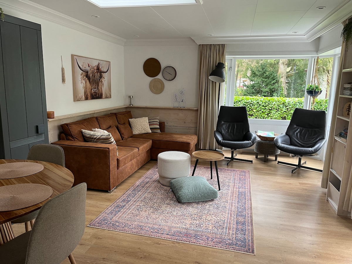 The living area features a spacious brown sofa arranged around a light area rug, complemented by two black armchairs. A small round table is included. Natural light fills the room from large windows, which offer a view of the greenery outside. Decorative wall art adds character.