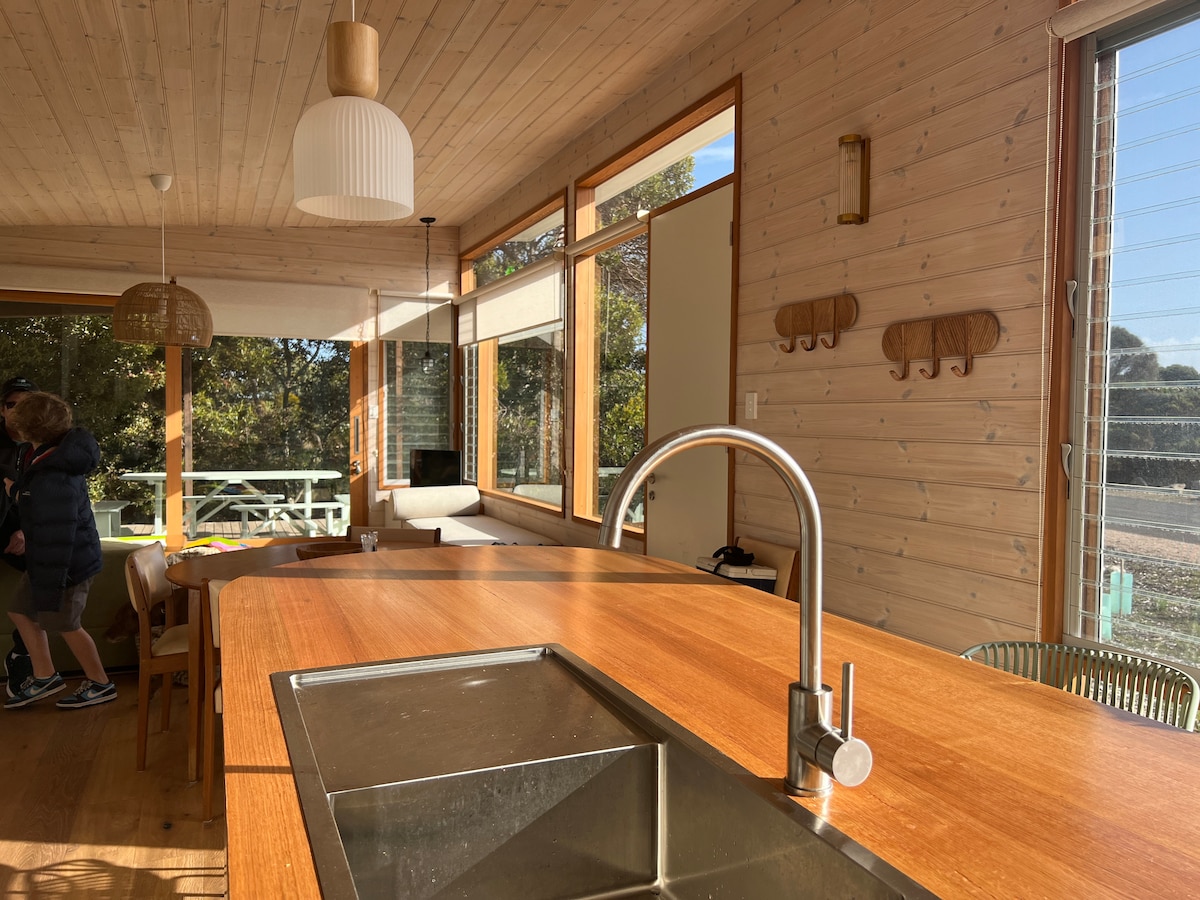 A contemporary kitchen area features a wooden island with a stainless steel sink, creating a functional space. Large windows allow natural light to fill the room, complemented by light wooden paneling. Simple decor and seating options are visible, enhancing the open-plan living concept.
