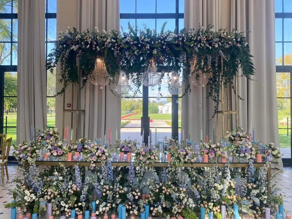 A tastefully arranged wedding ceremony backdrop features a floral display with a variety of pastel flowers and greenery. Elegant chandeliers hang from a floral arch above, while a table adorned with colorful candles and additional floral arrangements complements the serene indoor setting.