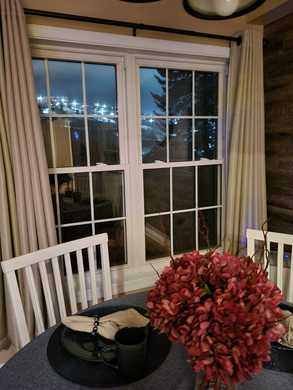 A dining area is featured with a round table set for two, accompanied by white chairs. A large window framed by light curtains allows for views outside, while a bouquet of vibrant red flowers adds a pop of color to the scene.