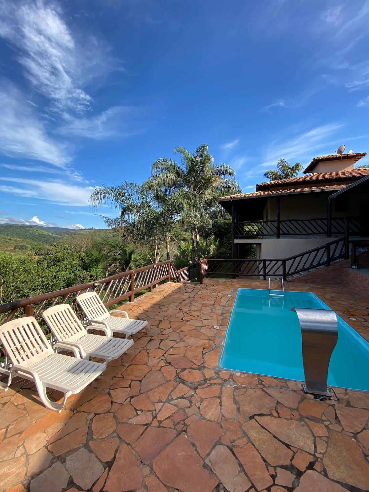 A private pool is set on a stone patio, surrounded by lush greenery. Four reclining lounge chairs are positioned for relaxation. The backdrop features a clear blue sky and distant mountains, creating a serene outdoor space.