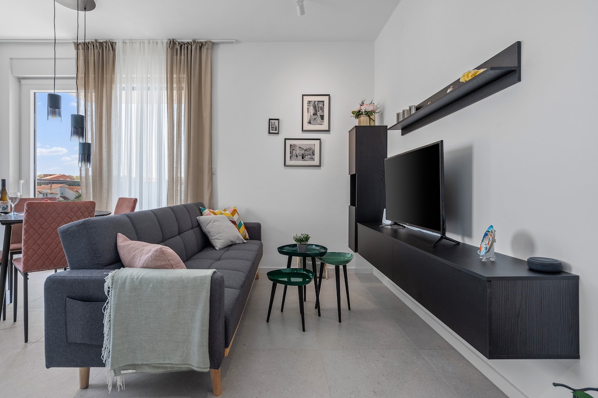 A modern living area features a gray sofa adorned with colorful cushions and an understated throw. Two green side tables are placed in front, with a mounted shelf and flat screen television on the adjacent wall. Natural light filters through the sheer curtains.