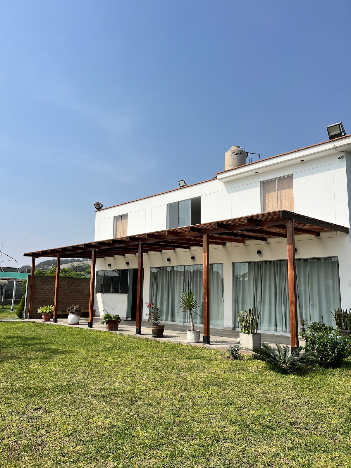 The house features a spacious patio area supported by wooden beams, with large glass windows offering natural light. Surrounding green gardens are visible, filled with various potted plants and a well-maintained lawn, creating an inviting outdoor environment.