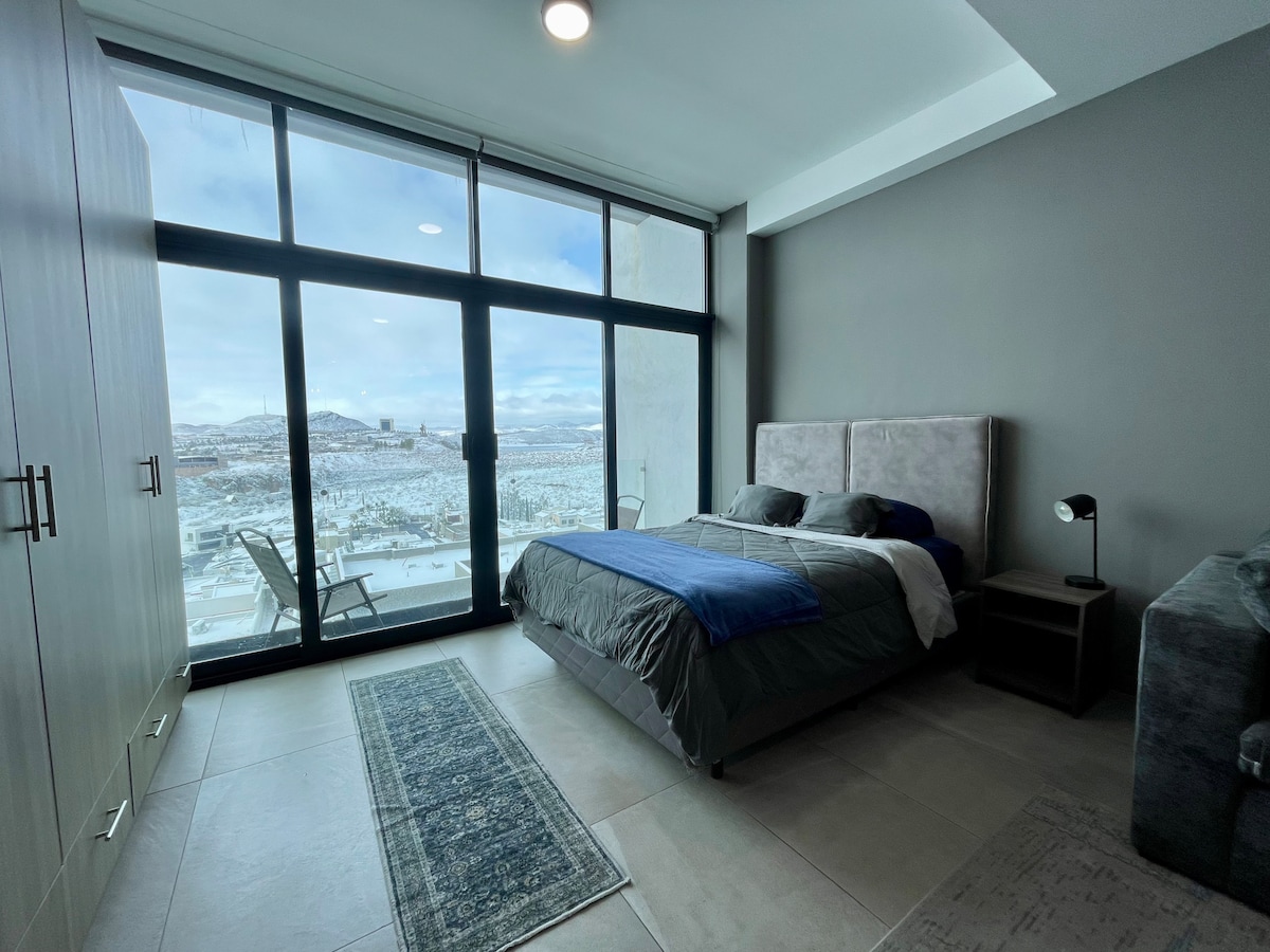 A spacious bedroom features large glass doors that provide a panoramic view of the surrounding landscape. A comfortable bed is dressed in muted tones, complemented by a stylish rug and sleek wardrobe. Natural light fills the room, enhancing the modern decor.