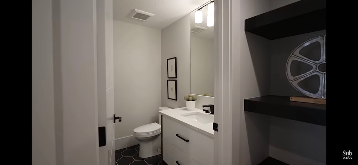 A modern bathroom features a white vanity with ample storage, complemented by a sleek mirror above. A minimalist décor is noted with framed artwork on the walls and a small potted plant. The floor showcases stylish hexagonal tiles, enhancing the contemporary feel.