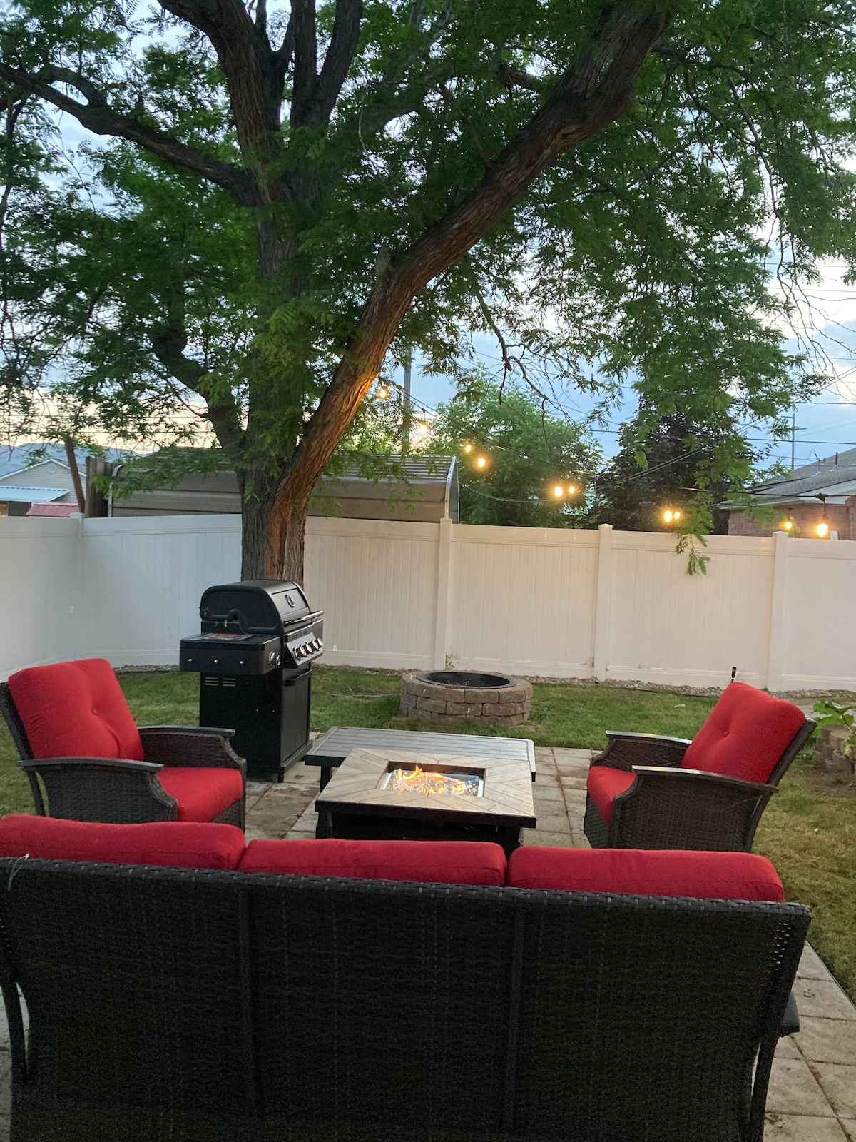 An outdoor seating area is furnished with four red cushioned chairs arranged around a stone fire pit. A barbecue grill is positioned nearby, and string lights are visible overhead, enhancing the evening ambiance. A grassy area is surrounded by a white privacy fence and a tree.
