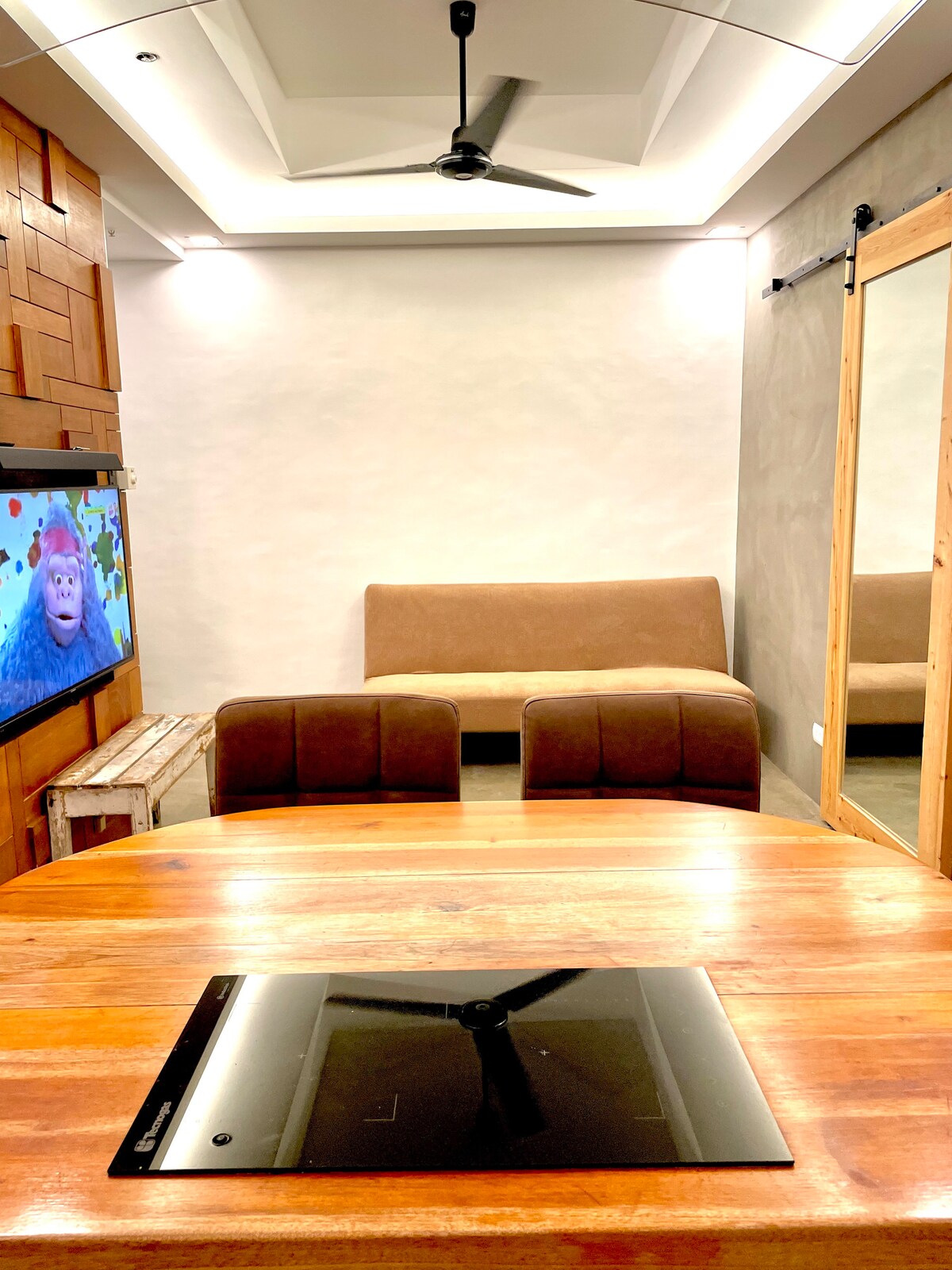 A living area features a cozy sofa bed and two upholstered chairs facing a television mounted on the wall. Natural light is provided through a window, and a wooden dining table with a stove is prominently displayed in the foreground.