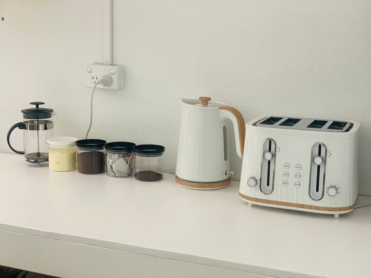 A simple kitchen countertop displays an electric kettle, a coffee maker, and a toaster. Containers hold coffee and sugar, ensuring essentials are within reach. The white and wood accents create a clean and inviting space for meal preparation and beverage crafting.