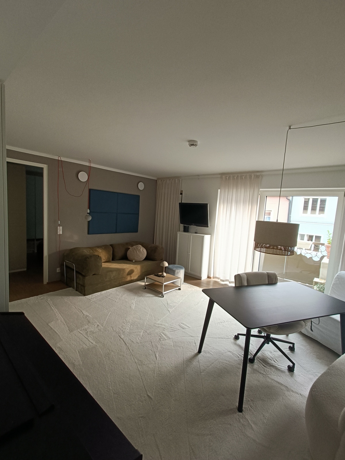 A modern living area is presented with a cozy sofa and a small side table. A desk with a rolling chair offers a workspace. Natural light filters through sheer curtains, while a television is mounted on the wall. Neutral colours and minimal decor create a calm environment.