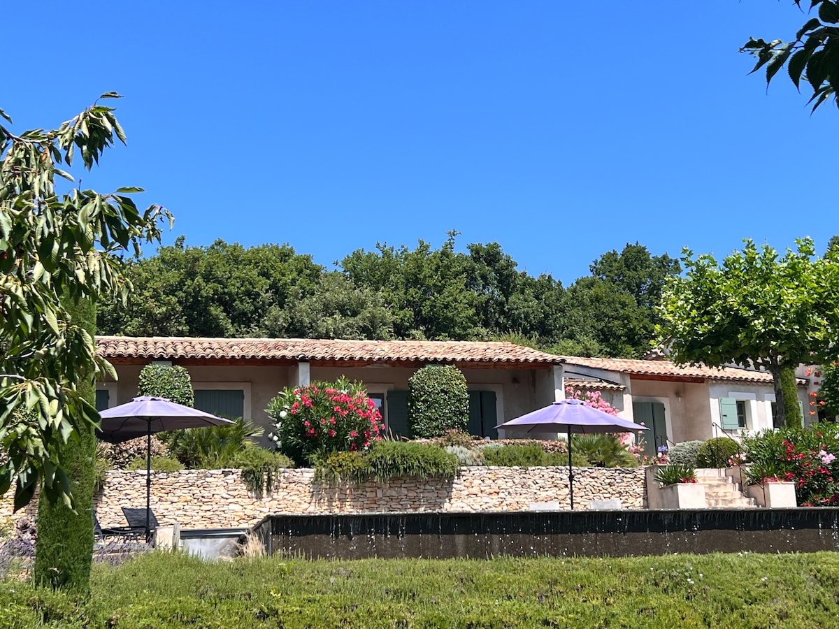 A charming cottage is surrounded by lush greenery and vibrant flowering plants. Two umbrellas offer shade in the outdoor area, while a stone wall borders a neatly maintained garden. The clear blue sky complements the tranquil setting, creating a serene atmosphere.