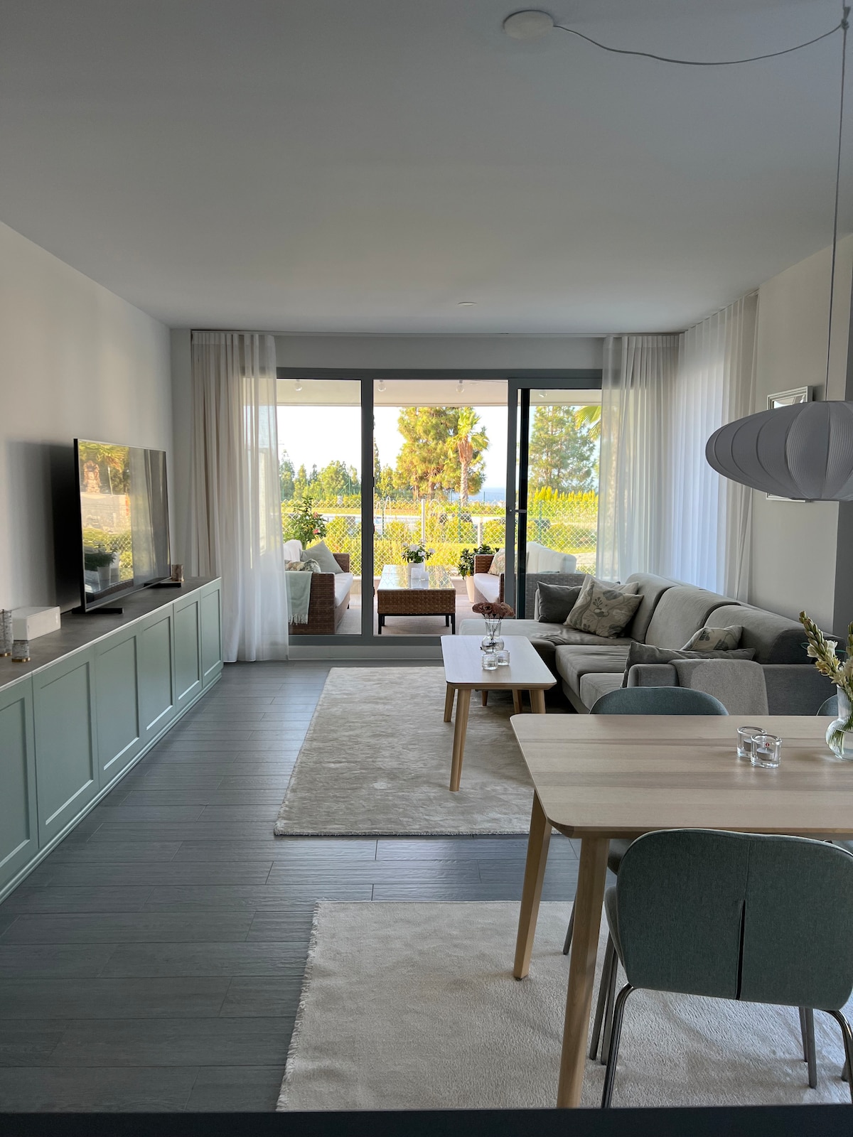 A modern living space features a comfortable seating area with a soft gray sofa, a dining table for four, and a large flat-screen TV. Floor-to-ceiling windows allow natural light to illuminate the room, offering views of greenery outside. Light curtains provide an airy feel.