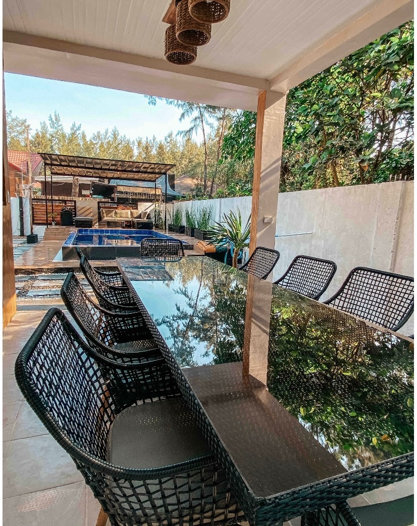 A spacious outdoor dining area features a long table made of glass, surrounded by black woven chairs. Lush greenery and a swimming pool are visible in the background, creating a tranquil setting for gatherings.