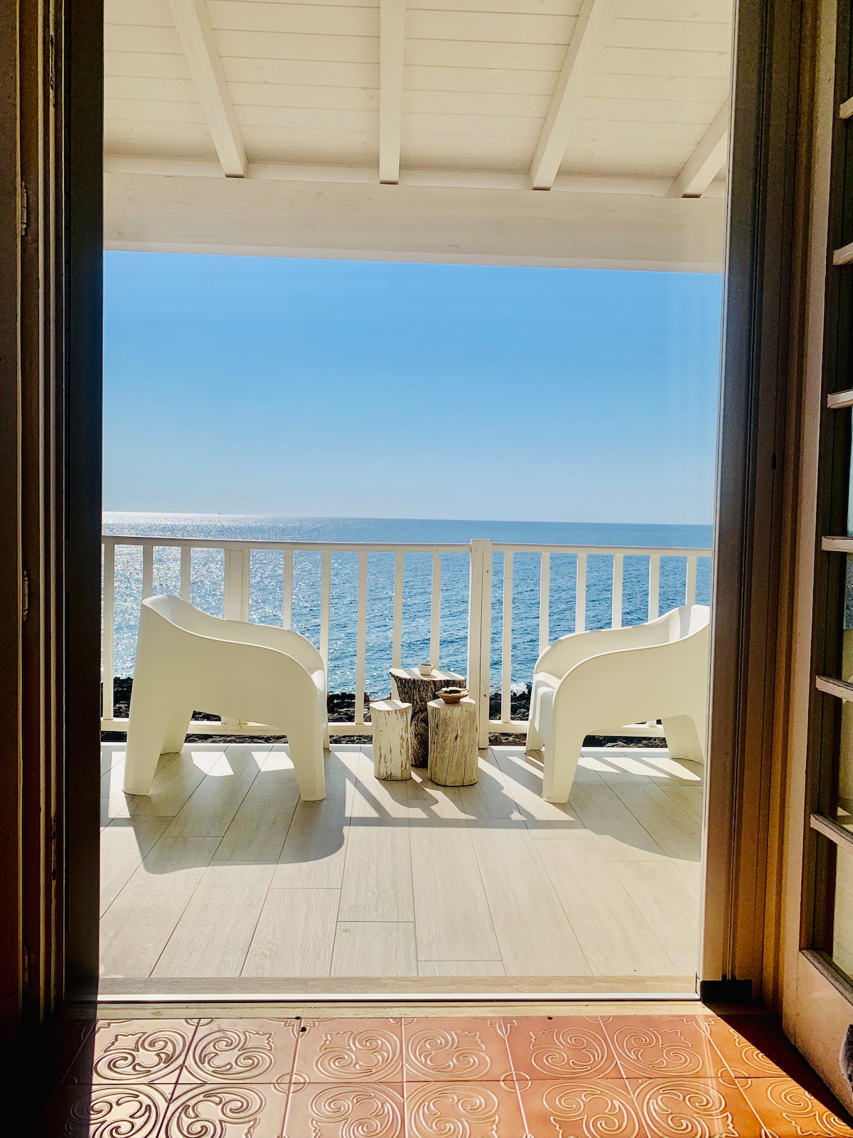 A serene view from the doorway reveals two white chairs positioned on a balcony, overlooking calm blue waters. A small wooden table stands between the chairs, inviting relaxation while enjoying the picturesque seaside scenery.