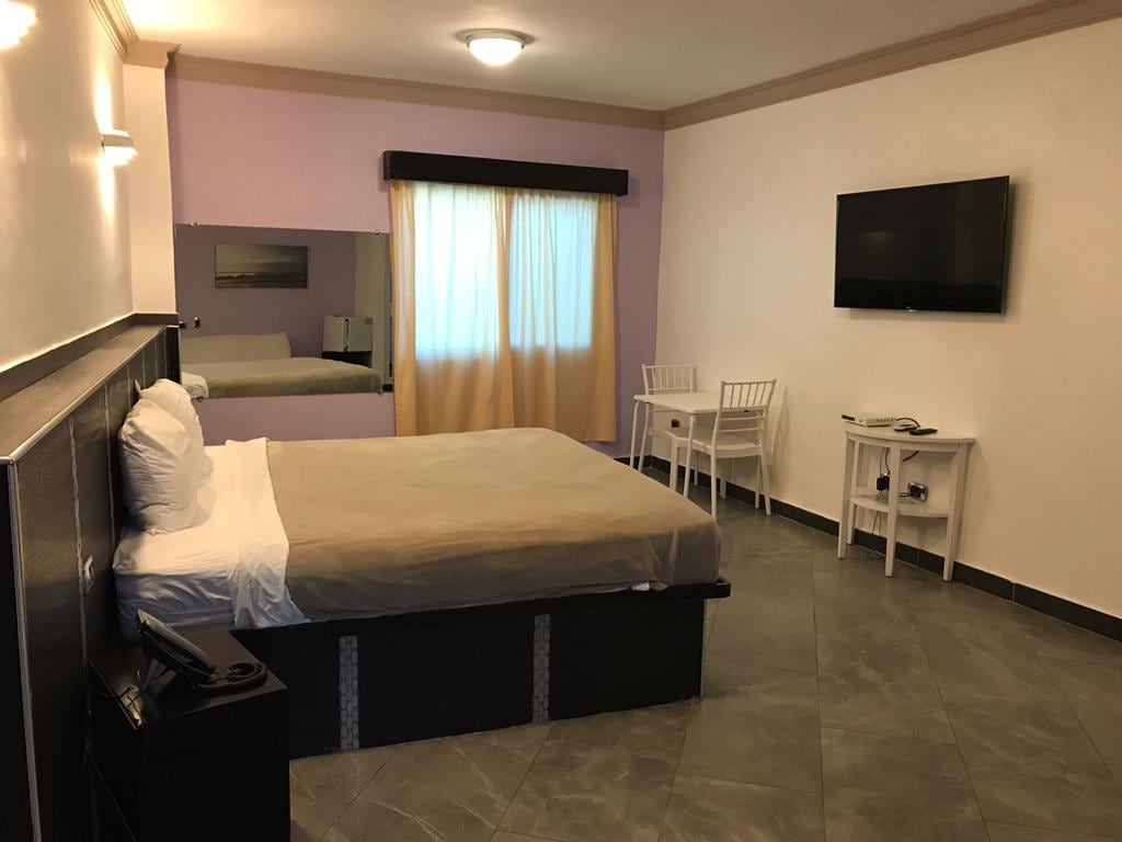 A well-lit room features a spacious bed with a neutral-colored coverlet. A flat-screen TV is mounted on the wall, and a small dining area is equipped with a table and two chairs. Natural light filters through the sheer curtains, creating a cozy atmosphere.