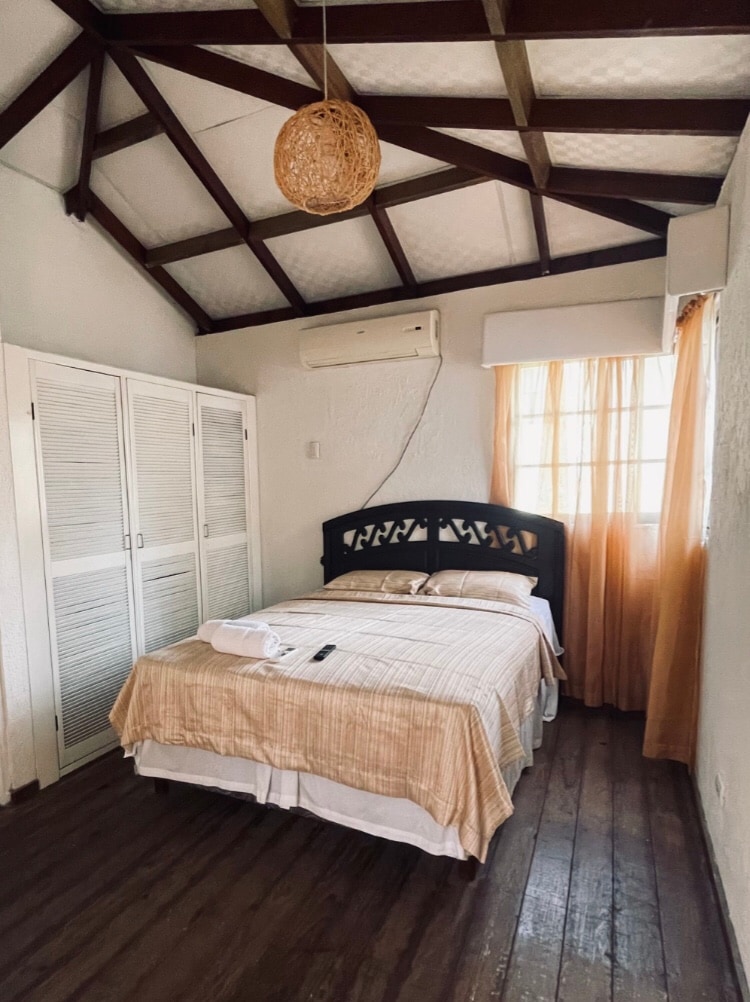 A cozy bedroom features a dark wooden bed frame with a light-colored bedspread. Natural light enters through a window adorned with sheer curtains. A ceiling fan and woven light fixture are present, and built-in closets provide storage against one wall.