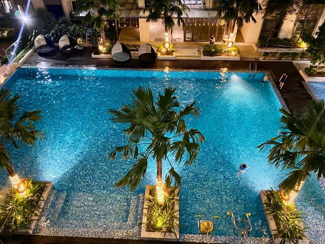 An expansive swimming pool is surrounded by lush greenery and palm trees. Soft lighting enhances the tranquil atmosphere, and lounge seating areas are placed around the pool's edge. The clear, turquoise water reflects ambient light, inviting relaxation.