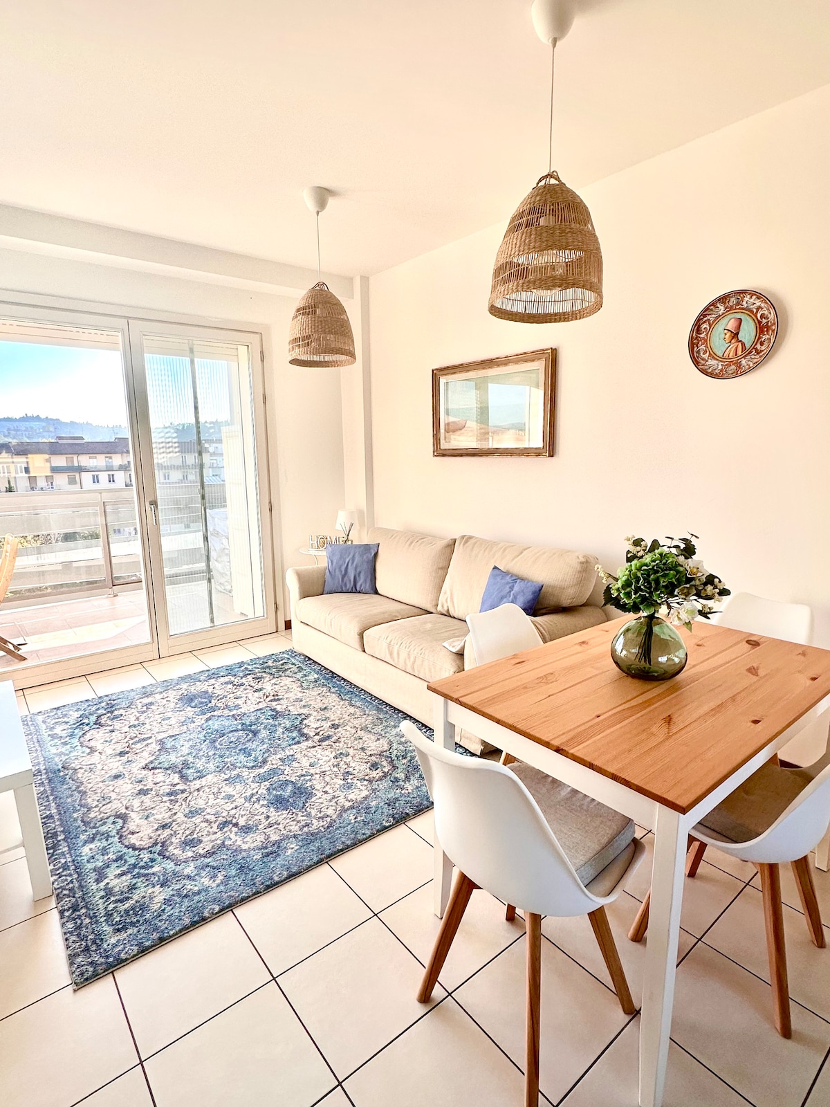 A well-lit living area features a comfortable sofa adorned with decorative cushions, complemented by a wooden dining table surrounded by white chairs. A stylish rug adds a touch of color, while large glass doors open to a balcony with city views.