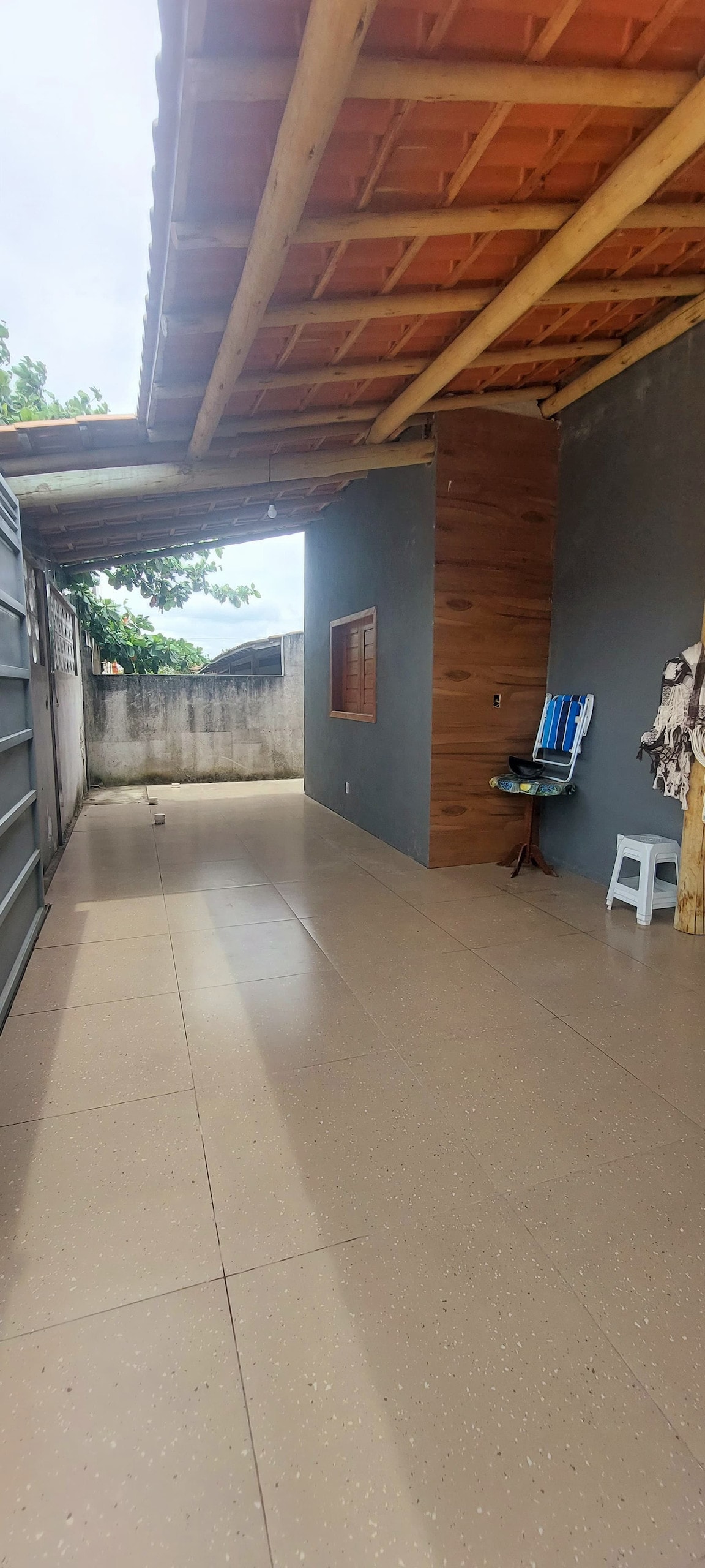 An outdoor area features a covered porch with a tiled floor and walls finished in neutral tones. A rustic wooden accent wall adds texture, while a small stool and a deck chair provide seating options. Natural light filters in, creating a bright and open space.