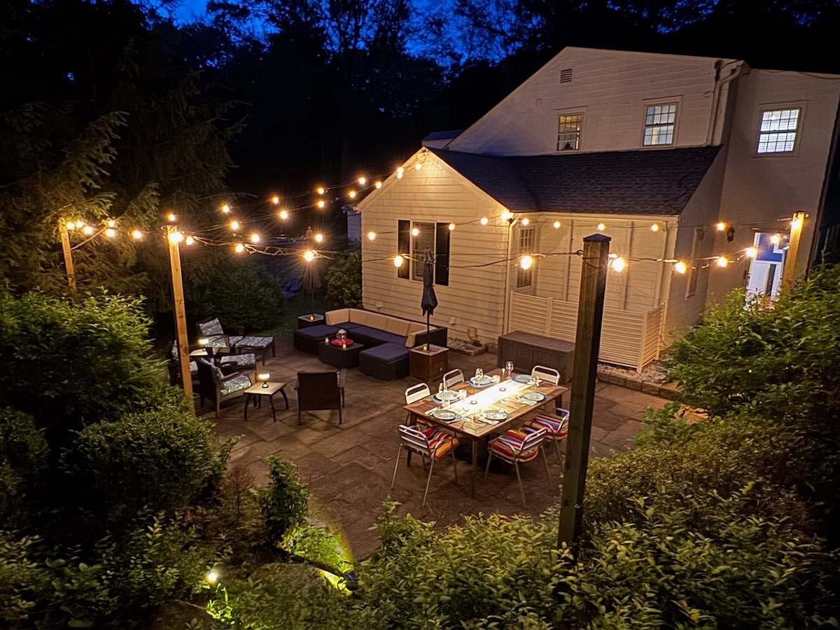 The outdoor dining area features a large table surrounded by colorful chairs, illuminated by string lights. A cozy seating area with a dark sectional sofa is visible nearby, set against the backdrop of the house, creating an inviting space for gatherings.