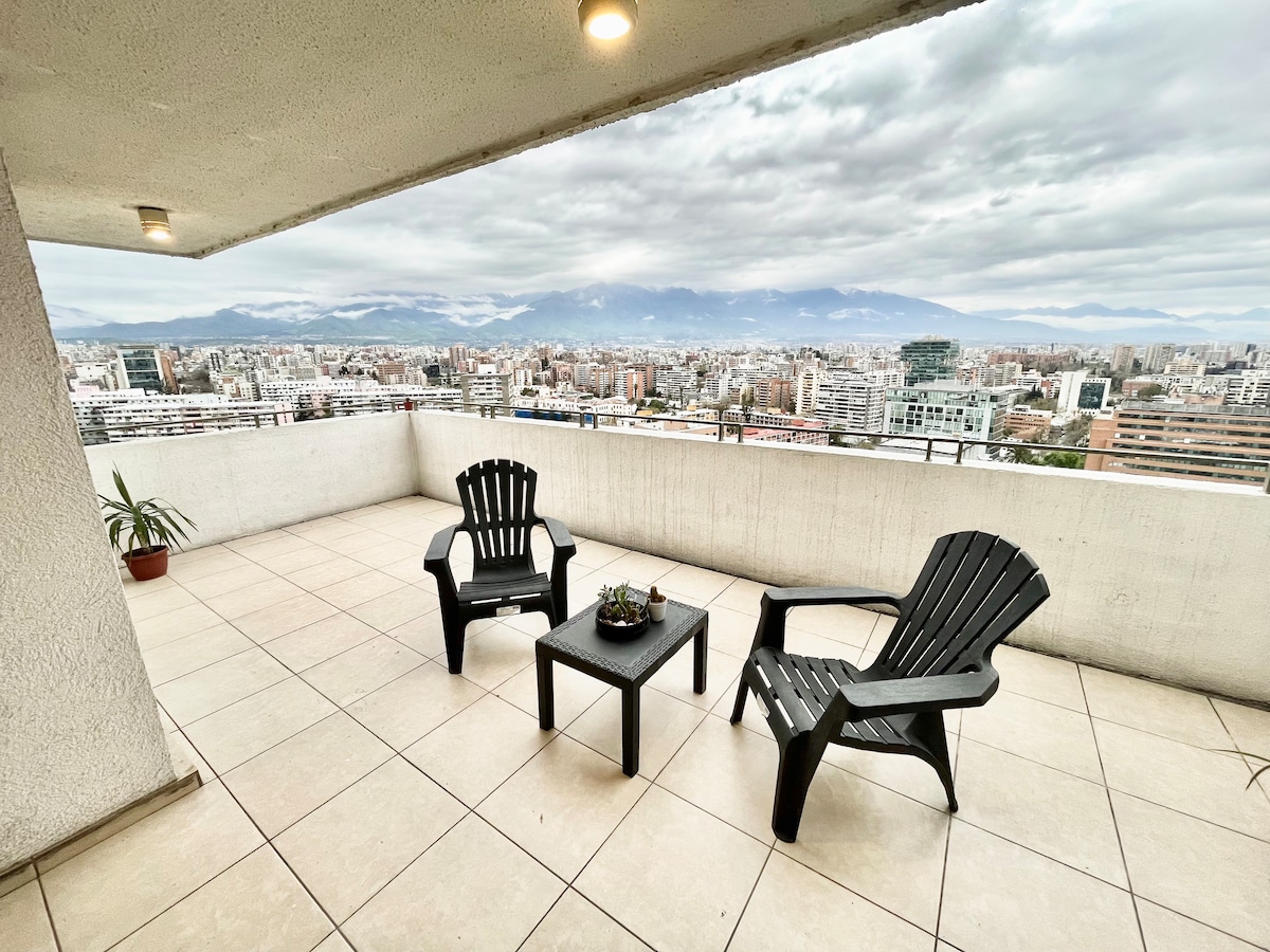 A spacious terrace features two black Adirondack chairs positioned around a small table. A potted plant is situated in the corner. The urban skyline extends into the distance, with mountains visible beneath a cloudy sky.
