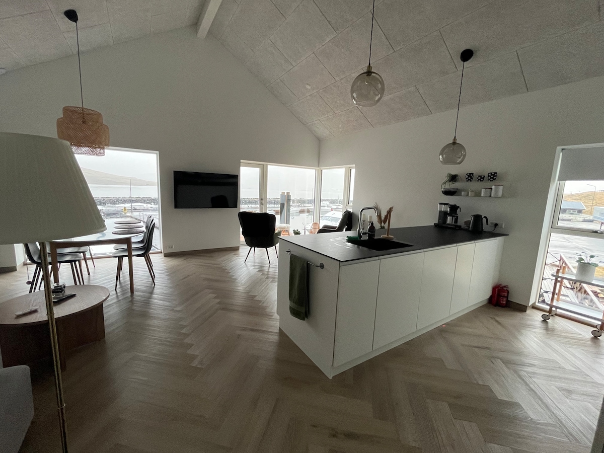 An open-concept kitchen and dining area is presented, featuring a central island with minimalist cabinetry. Large windows allow for natural light and offer views of the surrounding landscape. A comfortable seating arrangement is visible, alongside a wall-mounted television enhancing the modern design.