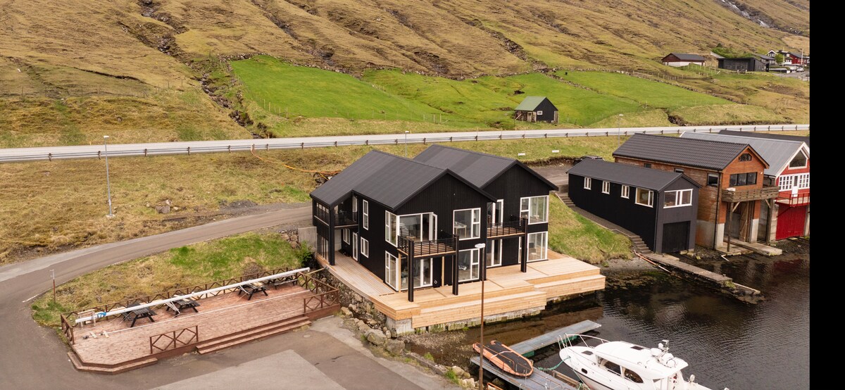 An aerial view displays a modern black house on the waterfront, surrounded by lush green hills. A deck extends toward the water, and several boats are docked nearby. The landscape is bordered by a paved road and additional buildings visible in the background.