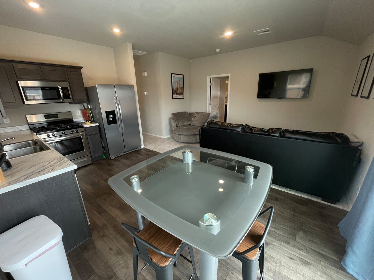 A modern kitchen is featured with stainless steel appliances and a marble countertop. A glass dining table with four chairs is positioned centrally. In the background, a cozy living area includes a large sectional sofa and a wall-mounted television, enhancing the communal space.