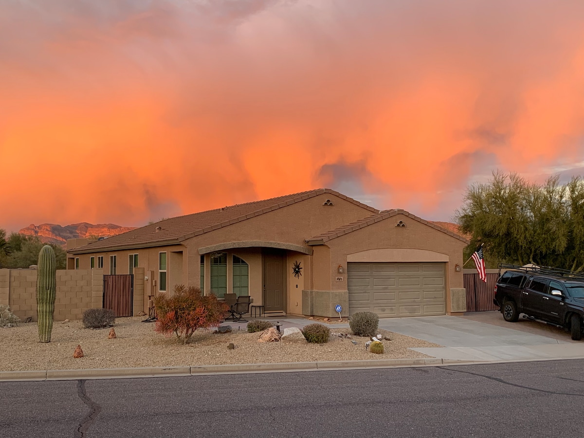 The home's exterior is showcased against a vibrant sunset, with hues of orange and pink in the sky. Desert landscaping surrounds the property, featuring cacti and shrubs. A driveway leads to a two-car garage, and a welcoming front porch offers seating for relaxation.