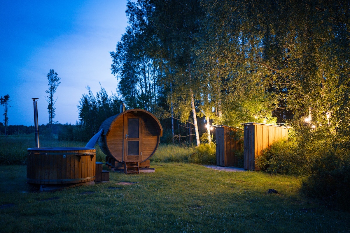 A serene outdoor space is observed, featuring a wooden hot tub beside a barrel-shaped sauna. Two showers and a toilet are placed a short distance away, illuminated by soft lighting among the greenery. The surrounding area is grassy and dotted with trees.