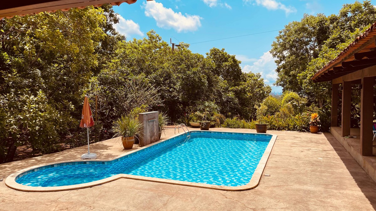 A large swimming pool is set in a spacious outdoor area, surrounded by lush greenery. The pool features a light blue tile design, and various potted plants are placed around the patio, enhancing the tranquil atmosphere.
