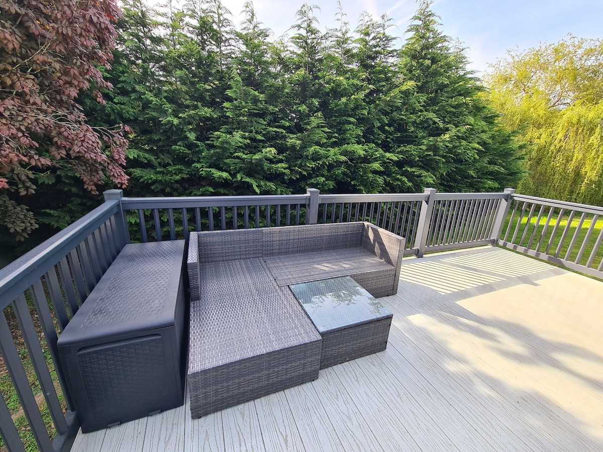 A spacious outdoor decking area features a modern rattan furniture set, including a sectional seating arrangement and a glass-topped coffee table. Lush green hedges provide privacy and natural beauty, while sunlight brightens the space.