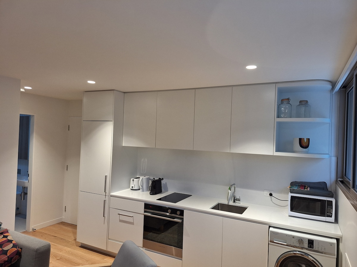 A modern kitchen space is equipped with sleek cabinetry and essential appliances, including a stove, oven, microwave, and refrigerator. Natural light fills the area through large windows. The countertops are finished in a smooth white surface, contributing to a clean and minimalist design.