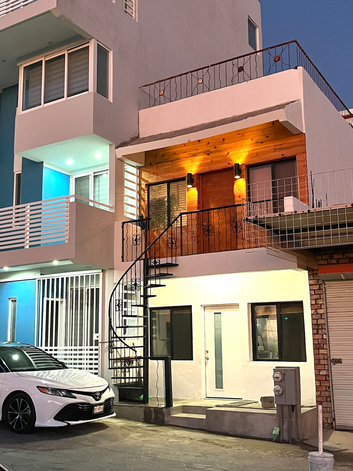 A welcoming entrance area showcases a spiral staircase leading to a wooden door, illuminated by warm lights. The façade features a combination of bright colors and clean lines. A parked car is visible on the left, while the street view includes surrounding buildings.