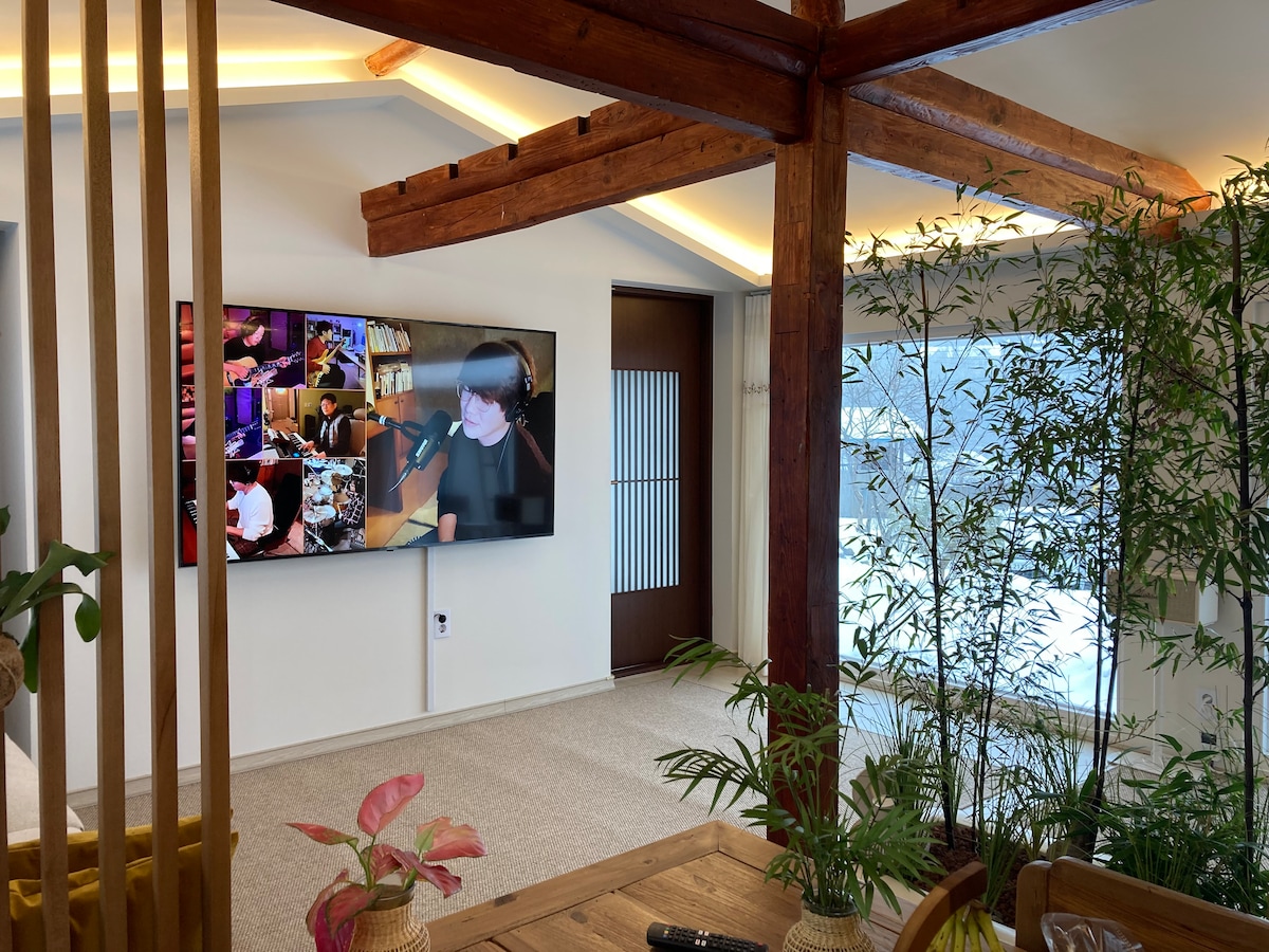 A spacious living area is highlighted by wooden beams and large windows that allow natural light to fill the room. A flat-screen TV is mounted on the wall, displaying vibrant images. Green plants are placed throughout, adding a touch of nature to the decor.