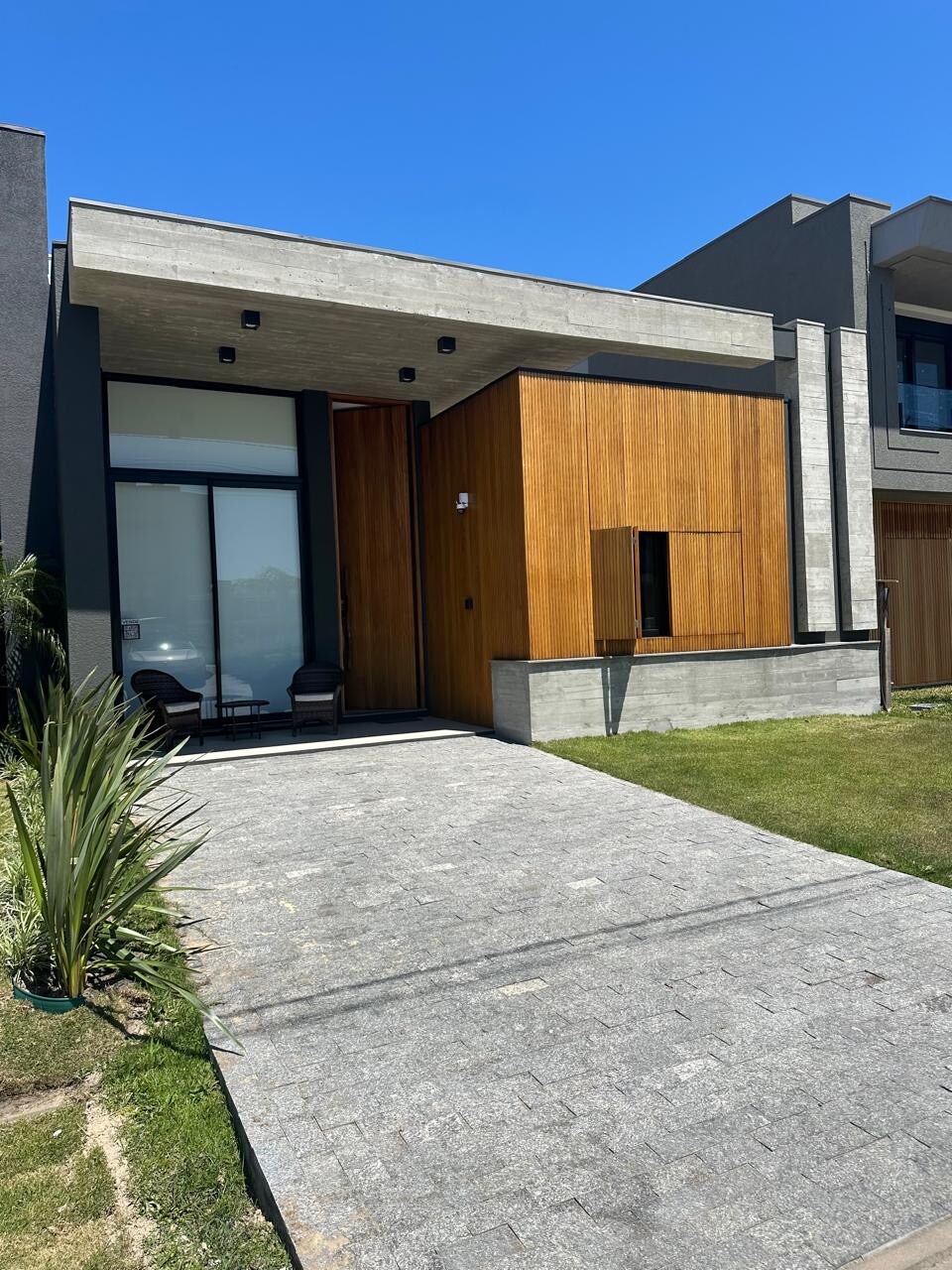 The exterior of a modern house is depicted, featuring a combination of stone and wood materials. A paved pathway leads up to the entrance, flanked by manicured grass. Two chairs are positioned near the front door, creating an inviting approach.