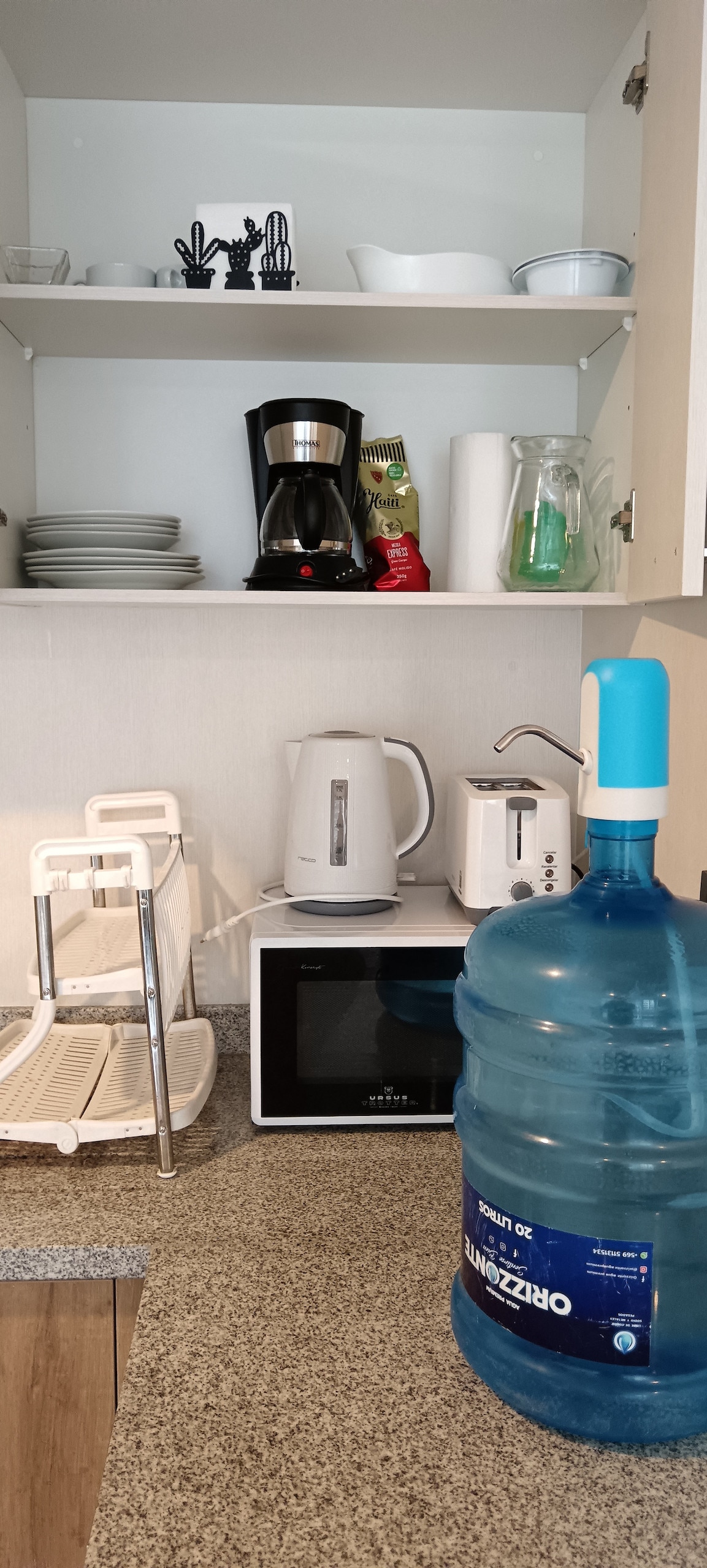 The kitchen area features a microwave and coffee maker on a granite countertop. White dishware is neatly arranged on shelves above, while a large water bottle and a kettle are also visible. This space is organized for convenience and functionality.