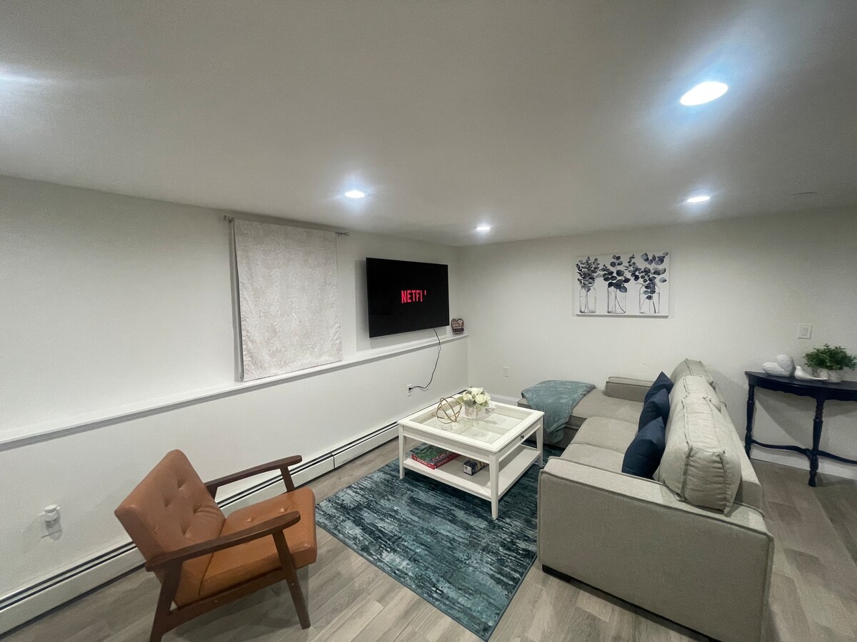 The living room features a comfortable sofa and a stylish armchair situated on a blue rug. A coffee table sits at the center, with a large wall-mounted Smart TV displaying the Netflix logo. Soft lighting enhances the overall ambiance.