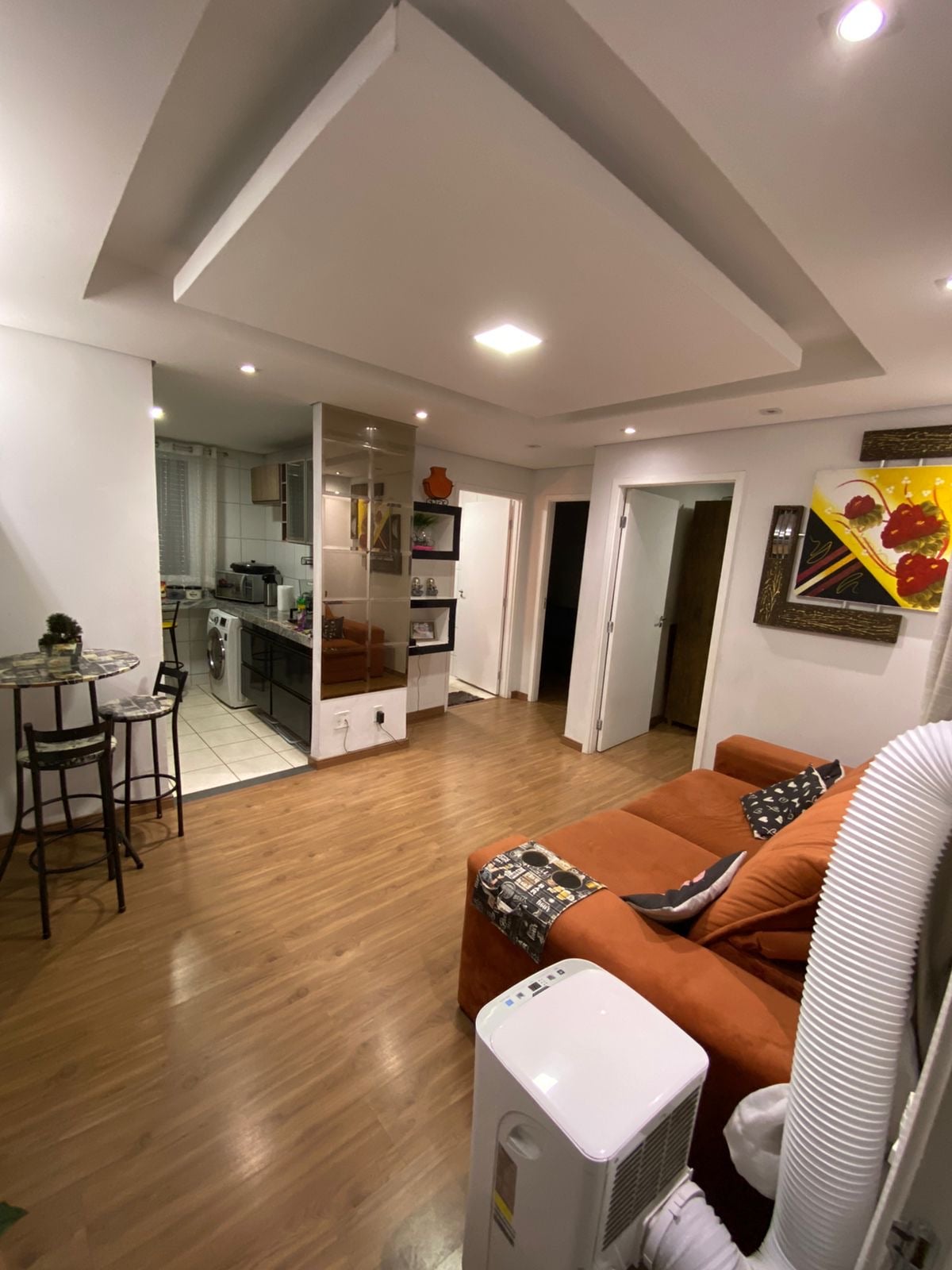 An open living space features a comfortable orange sofa facing a wall adorned with vibrant artwork. A small dining area with two chairs and a table is visible, leading to a well-equipped kitchen with modern appliances. Soft lighting enhances the room's inviting atmosphere.