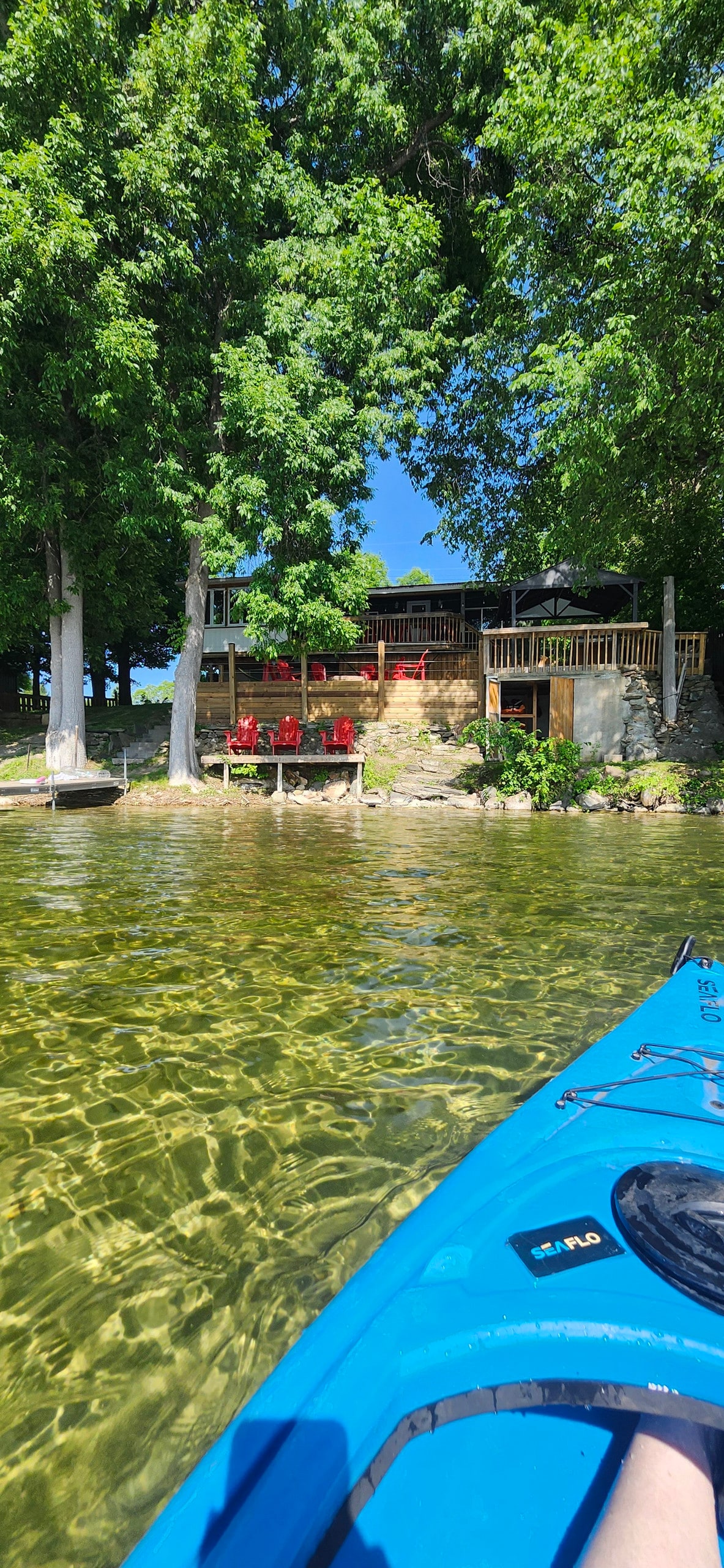 The view from the water captures the cottage nestled among lush green trees. A spacious deck with red chairs overlooks the lake. The clear water reflects the bright sky, inviting guests to enjoy the serene surroundings.