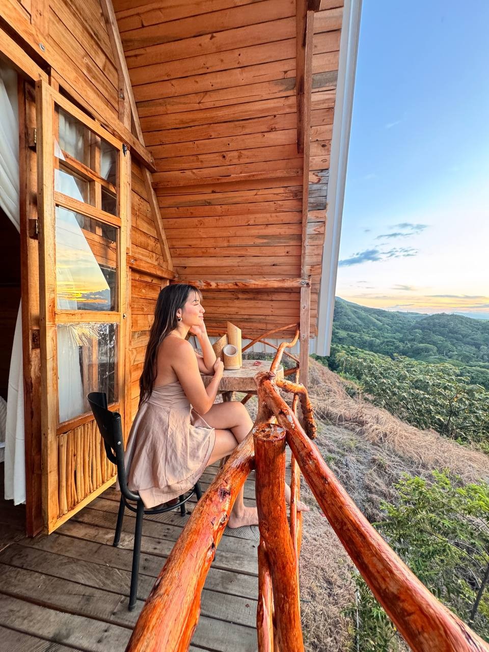 A wooden deck offers a serene view of rolling hills and a colorful sunset. A small table and chair are placed on the balcony, creating a perfect spot for relaxation. The rustic wooden railing enhances the connection with nature.