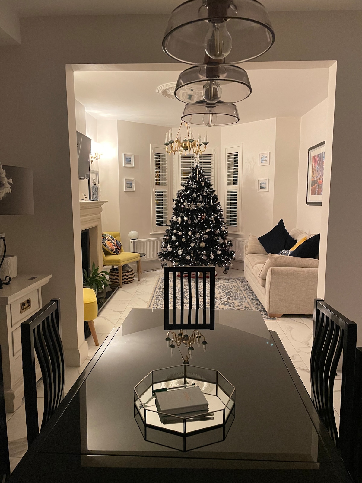 A modern dining area is presented with a sleek black table and four chairs. A decorative centerpiece is displayed on the table. In the background, a cozy living room is visible, featuring a white sofa and a Christmas tree, decorated with lights and ornaments.