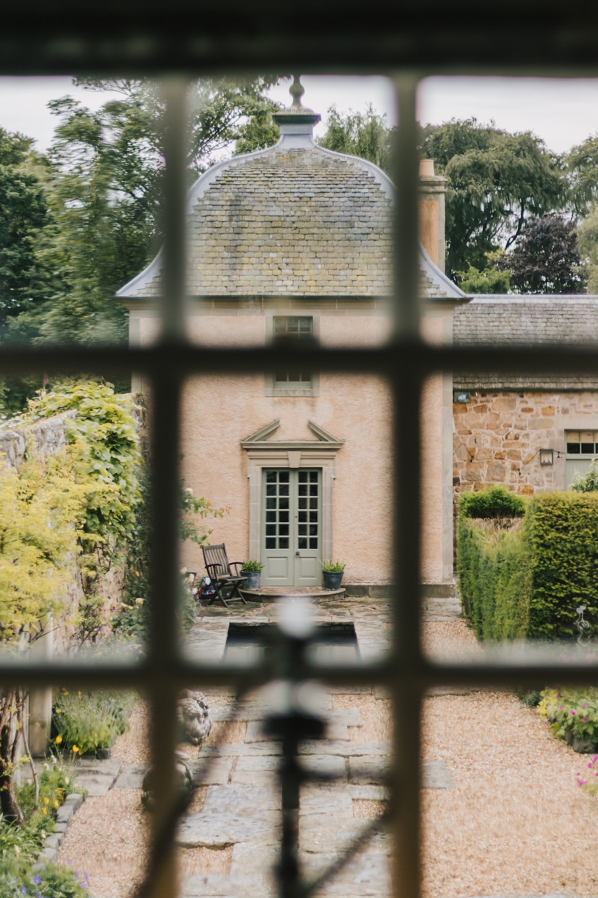 A view through a grid of an exterior door reveals a charming, pale pink building with a distinctive peaked roof. Lush greenery and a stone pathway lead to the entrance, while a chair on the patio invites relaxation in the tranquil garden setting.