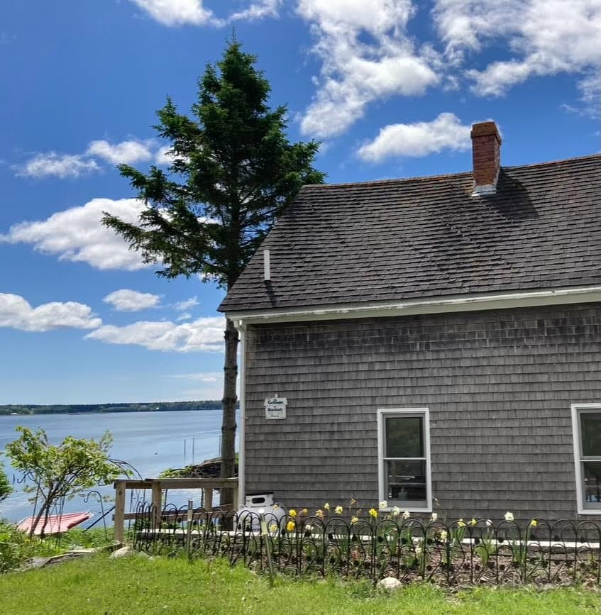 The exterior of Shackford Cove Cottage is shown, featuring gray shingles and a small deck area. A tree is seen beside the structure, with blooming flowers in a garden bed in the foreground. The tranquil waters of Shackford Cove and blue sky with clouds are visible in the background.